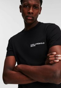 Crna pamučna majica kratkih rukava, s bijelim izvezenim logotipom "KARL LAGERFELD JEANS" na lijevoj strani prsnog područja.