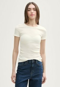 Krémový top s krátkým rukávem a strukturovaným, řaseným designem. Nosený s modrými džíny s vysokým pasem. Minimalistický styl, hladká látka.