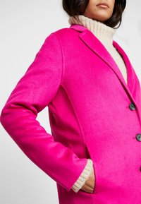Cappotto di lana di un vivace rosa con un design sartoriale, revers a punta, due bottoni e tasche laterali, sovrapposto a un maglione a collo alto color crema.
