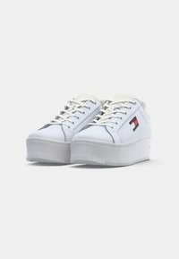 Weiße Plattform-Sneaker mit einer glatten Lederoberfläche, grauer Gummisohle, kontrastierendem blauem und rotem Logo-Patch an der Seite und weißen Schnürsenkeln.