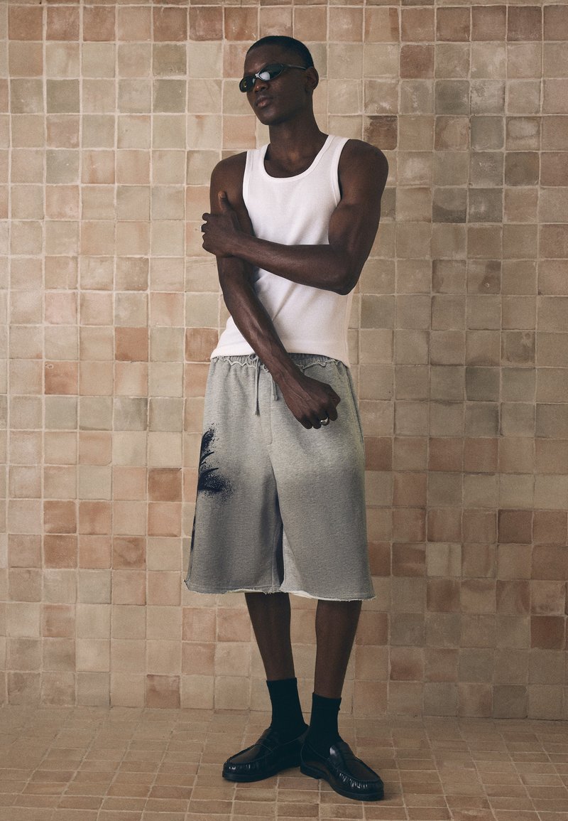 Homme portant des lunettes de soleil noires, un débardeur blanc, un short gris avec un motif noir, des chaussettes noires et des chaussures noires se tient contre un mur carrelé.