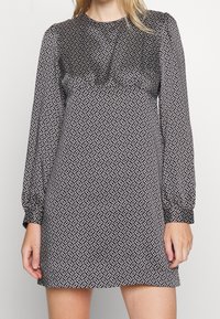 Černo-bílá vzorovaná šaty s dlouhými rukávy, kulatým výstřihem a geometrickým designem. Měkká tkanina, volný střih, nařasené detaily na dekoltu.