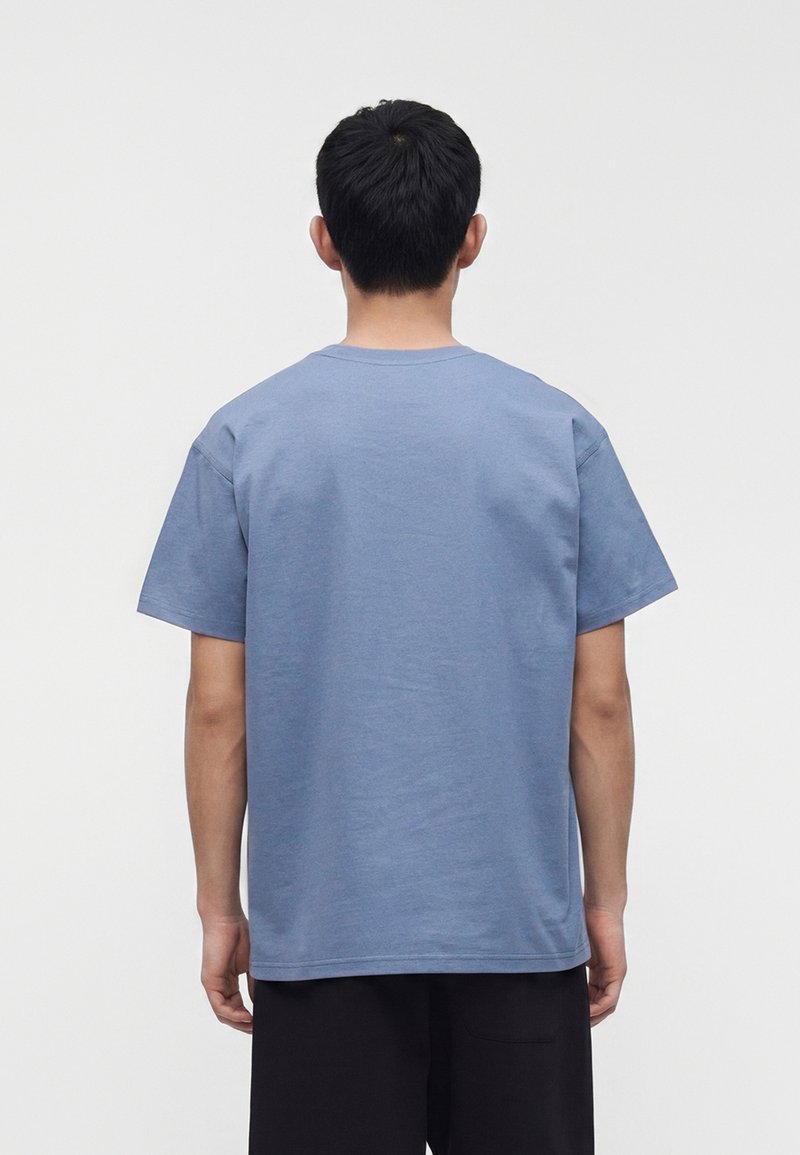 Man with short black hair wearing a plain blue short-sleeve shirt and black pants, seen from behind against a white background.
