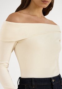 Nő krémszínű, vállat szabadon hagyó, bordázott felsőt visel, sötétkék farmerrel, amely a törzs közepénél van levágva.