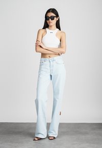 Bela brezrokavnik s potiskom "OFF", svetlo modri široki kavbojki z aplikacijo in črna sončna očala, stoji na nevtralni ozadju.