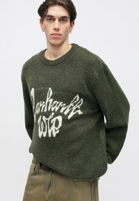 Pulover verde khaki cu o textură moale și un croi lejer. Are un design grafic alb pe față și un guler clasic la baza gâtului.