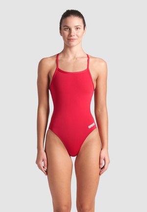 Femme debout et de face, portant un maillot de bain rouge une pièce à fines bretelles avec logo "arena" sur la hanche, fond neutre.