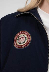 Gros plan sur une veste bleu marine avec une fermeture éclair argentée et un écusson brodé circulaire sur la poitrine portant l'inscription « Prohibited Motorsport Company Berlin Germany ».