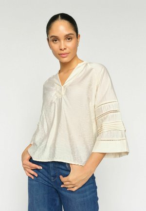 Frau trägt elfenbeinfarbene Bluse mit weiten Ärmeln, verziert mit Spitze und Plissees, kombiniert mit blauer Jeans, posiert vor einfarbig hellem Hintergrund.