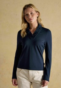 Mornarsko plavi dolgi rokav s volanastim V-izrezom in gladko teksturo; kombiniran s svetlimi hlačami, ki prikazujejo prileganje silhuete.