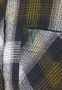 Gewebter Stoff mit einem karierten Muster in Marineblau, Creme und Olivgrün. Mit gesticktem "JV"-Logo in Türkis. Strukturierte Oberfläche.