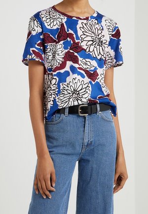 Personne portant une chemise à manches courtes avec de grandes fleurs blanches et des feuilles sur fond bleu et marron, associée à un jean bleu et une ceinture noire.
