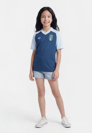 Jauna mergina šypsosi, dėvi mėlyną ir šviesiai mėlyną Italijos futbolo marškinėlį, džinsinius šortus ir baltus sportbačius, stovi priešais paprastą foną.