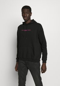 Black hooded sweatshirt made of cotton blend with a front pocket, featuring pink text "ONE COMMUNITY - DIVERSITY MATTERS." Soft texture.
