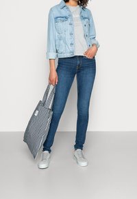 Light blue denim jacket, gray t-shirt with logo, dark blue skinny jeans, and striped tote bag. Light gray sneakers with thick soles.