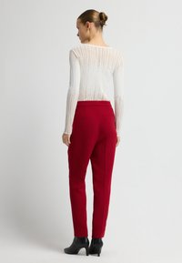 Frau mit einem Dutt, die ein weißes, strukturiertes Langarmoberteil, rote, hoch taillierte, maßgeschneiderte Hose und schwarze, hohe Stiefeletten trägt, mit dem Rücken zur Kamera.