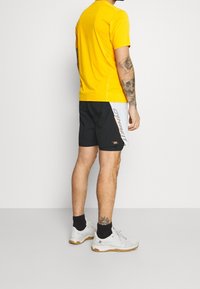 T-shirt de performance jaune, shorts noirs avec des accents blancs et orange, chaussures de sport blanches et chaussettes noires. La peau est visible au niveau des shorts.