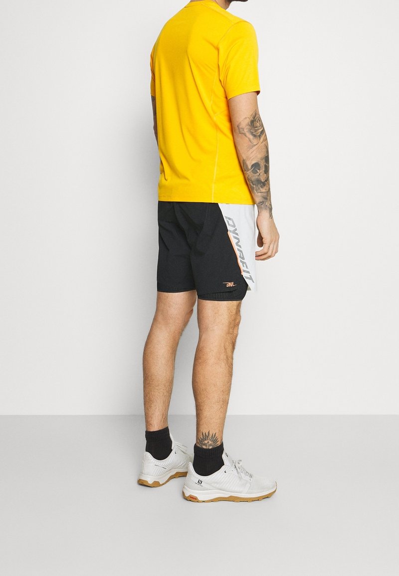 T-shirt de performance jaune, shorts noirs avec des accents blancs et orange, chaussures de sport blanches et chaussettes noires. La peau est visible au niveau des shorts.