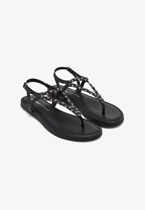 Sandalias negras con suela plana, que presentan una tira superior trenzada adornada con pequeños acentos metálicos. Diseño de talón abierto y con correa entre los dedos.