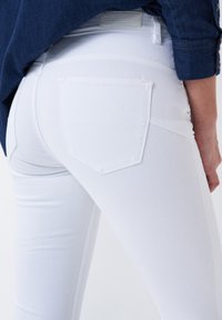 Jeans de mezclilla blanca con un diseño ajustado, que cuentan con bolsillos traseros y costuras visibles. Combinados con una blusa de mezclilla azul.