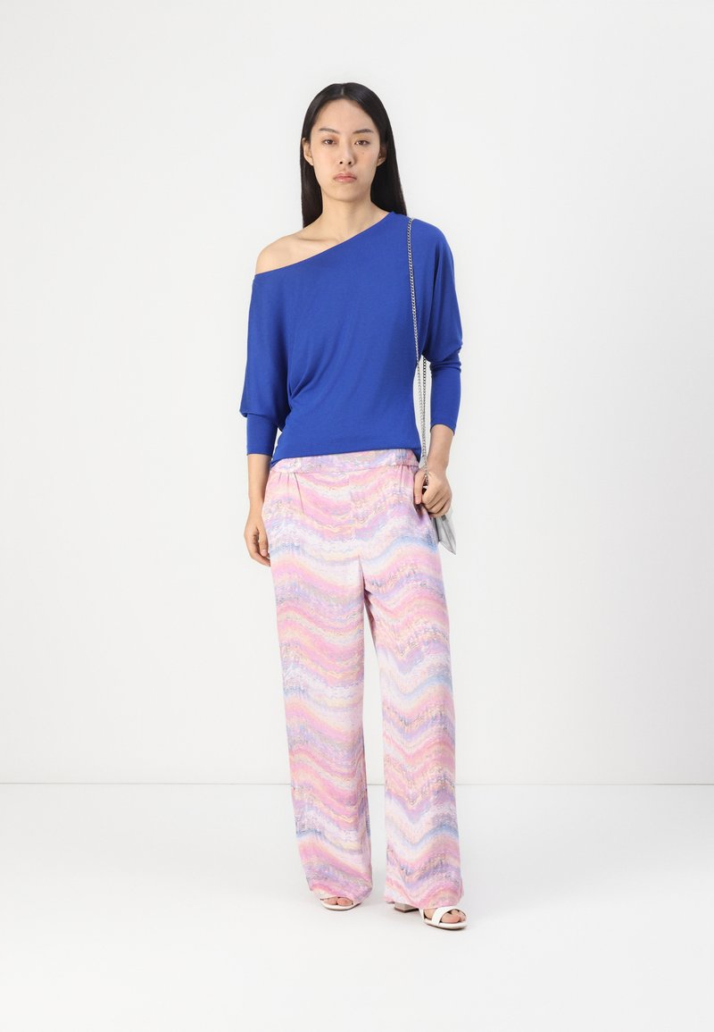 Blaues Off-Shoulder-Oberteil mit Dreiviertelärmeln, kombiniert mit pastellfarbenen pantaloons in Rosa, Blau und Weiß mit Muster. Schlichte weiße Sandalen.