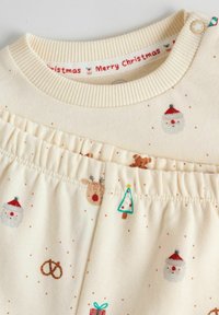 Pyjama bébé de couleur crème avec taille élastique et imprimés de visages du Père Noël, rennes, sapins de Noël, bretzels et texte "Merry Christmas" à l'intérieur du col.