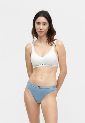 Woman with long dark hair wearing white Tommy Hilfiger bralette and blue underwear, standing against a plain light background.