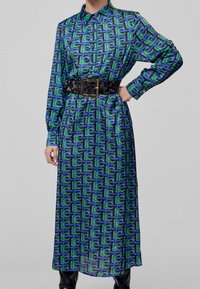 Långärmad klänning med krage i ett blått och grönt geometriskt mönster. Bärs med ett svart bälte som har en guldfärgad spänne. Slät, flödande tyg.