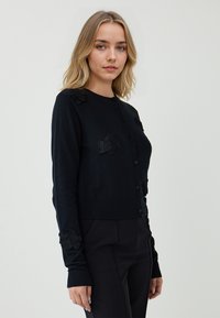 Schwarze Strickjacke mit langen Ärmeln, gerippten Bündchen und floraler Stickerei auf den Schultern. Mit rundem Halsausschnitt und vorderem Knopfverschluss.