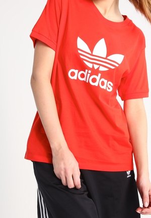 Osoba nosząca czerwoną koszulkę Adidas z białym logo oraz czarne spodnie Adidas z białymi paskami i logo na lewym udzie.