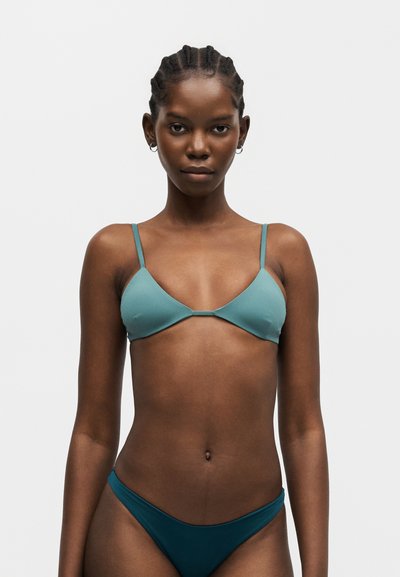 Jeune femme aux cheveux tressés portant un haut de bikini turquoise et un bas de bikini turquoise foncé, debout devant un fond clair uni.