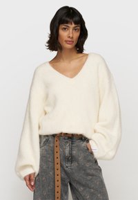 Weißer, flauschiger Strickpullover mit tiefem V-Ausschnitt und Ballonärmeln, kombiniert mit hoch-taillierten grauen gemusterten Jeans und einem braunen Gürtel.