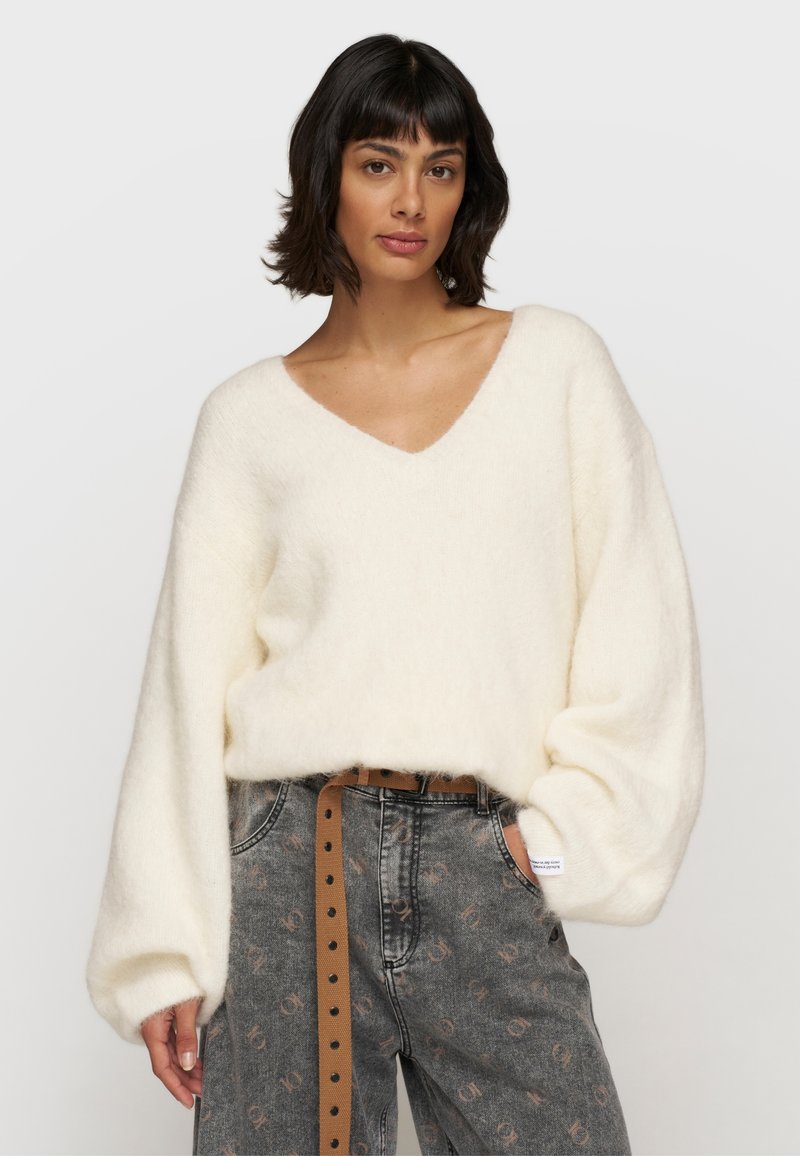 Weißer, flauschiger Strickpullover mit tiefem V-Ausschnitt und Ballonärmeln, kombiniert mit hoch-taillierten grauen gemusterten Jeans und einem braunen Gürtel.