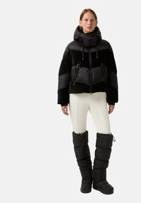 Sort, quilted pufferjakke med høj krave og snoretræk, parret med hvide tætsiddende bukser og sorte polstrede benvarmere.