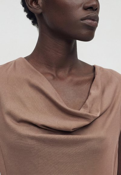 Κοντινό πλάνο ενός ατόμου που φοράει ένα ανοιχτό καφέ αμάνικο τοπ με δίπλωμα, εμφανίζοντας τον λαιμό, τη γραμμή του σαγονιού και μέρος του άνω στήθους.