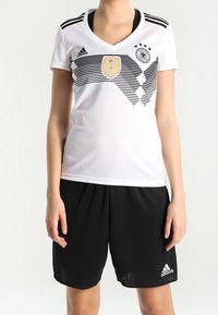 Bílý sportovní dres Adidas s černými pruhy a zlatým emblémem, doplněný černými šortkami s logem Adidas.