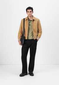Une veste bomber marron clair sur un polo vert, avec un col boutonné. Un pantalon cargo noir et des chaussures noires. Un sac à bandoulière noir minimaliste.