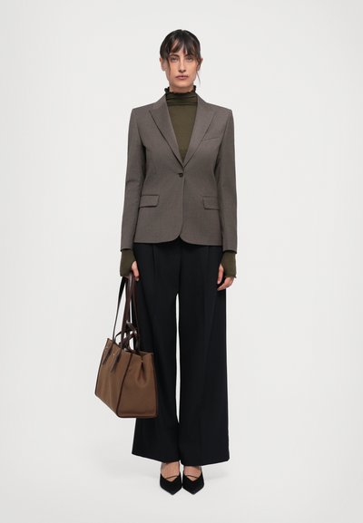 Brūns pielāgots žakets virs olīvu krāsas turtlenecka, apvienots ar melniem plašiem biksēm. Modelis tur brūnu rokassomu un valkā melnus šaurus apavus.