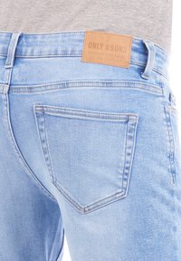 Jeans de mezclilla azul claro con una etiqueta de cuero marrón que lleva el texto "ONLY & SONS". La tela cuenta con un diseño clásico de cinco bolsillos y costuras visibles.