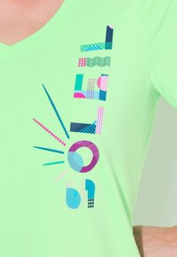 V-hals T-shirt i lysegrønt stof med flerfarvede geometriske mønstre og abstrakte former i pink, blå og mørkegrøn.