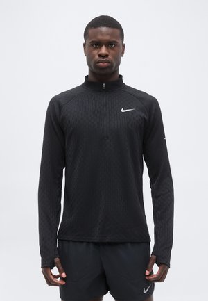 Uomo che indossa una maglia nera Nike con maniche lunghe e zip e pantaloncini neri Nike, in piedi rivolto in avanti su uno sfondo bianco semplice.