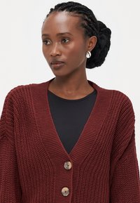 Burgunderstrikket cardigan med v-hals, med tre knapper, brede ermer og et teksturert mønster, parret med en svart topp under.