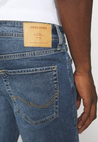 Jack & Jones -denimfarkut, joissa keskitummansininen pesu, keltaiset tikkauslangat ja takana nahkainen merkki, jossa on brändin logo.
