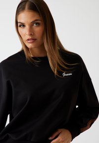 Felpa nera con scollo a girocollo, che presenta un logo bianco "Guess", maniche lunghe e righe marroni texturizzate lungo le braccia.