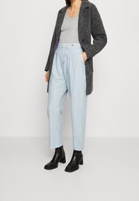 Grauer strukturierter Mantel über einem hellgrauen Oberteil, kombiniert mit hochgeschnittenen hellblauen Hosen und schwarzen Stiefeletten mit quadrater Zehenform.