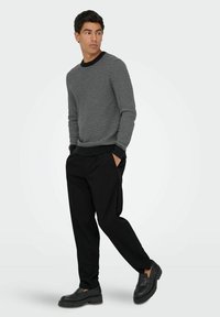 Schwarz-weißer gemusterter Pullover mit gerippten Bündchen, kombiniert mit lockeren schwarzen Hosen und schwarzen Schuhen, die ein modernes Design präsentieren.