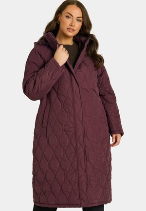 Femme portant un long manteau matelassé violet foncé avec un col montant, tenant le col d'une main et se tenant devant un fond uni.