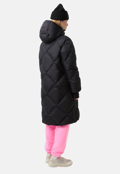 Zwarte pufferjas met een geruit diamantpatroon, capuchon en lange mouwen. Draagt over roze joggingbroek en beige sneakers.