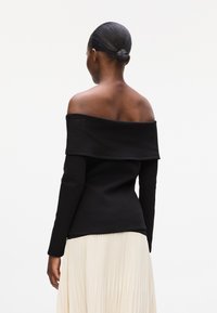 Black off-shoulder top with long sleeves, fitted design, and soft texture, worn with a cream pleated skirt.
