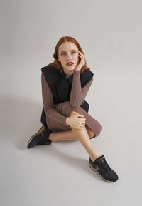 Schwarze sportliche Sneakers aus strukturiertem Material, kombiniert mit einem leicht braunen, langärmeligen Kleid mit Rippstruktur. Das Model sitzt lässig vor einem neutralen Hintergrund.